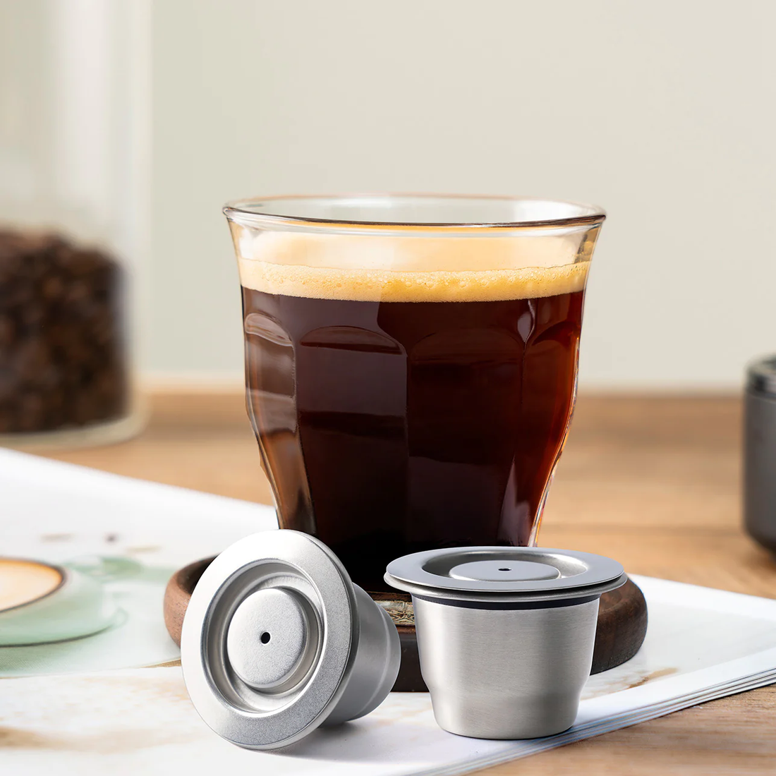 Two stainless steel reusable coffee capsules beside a transparent espresso glass filled with espresso, showcasing a cozy kitchen counter setting